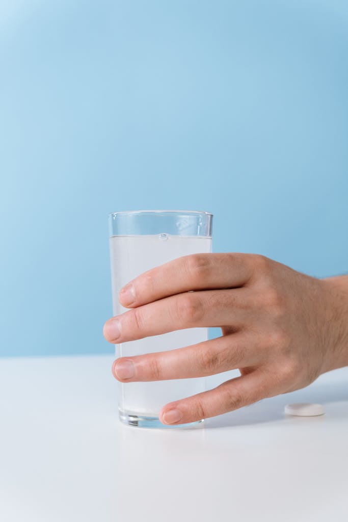 A hand holding a glass of effervescent water with a tablet on a white table.