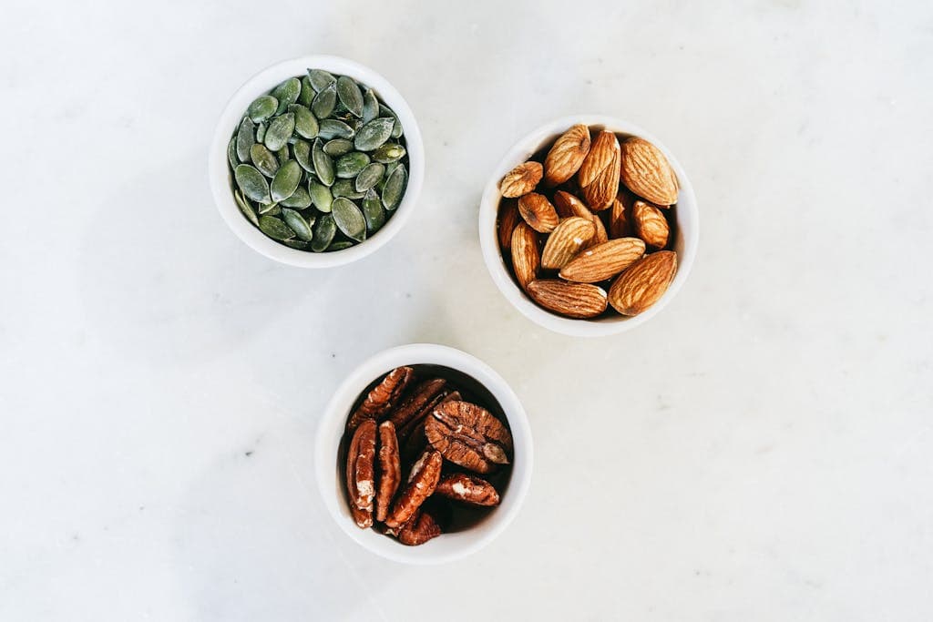 A top-down view of assorted nuts in bowls, featuring almonds, pumpkin seeds, and pecans, on a marble surface.