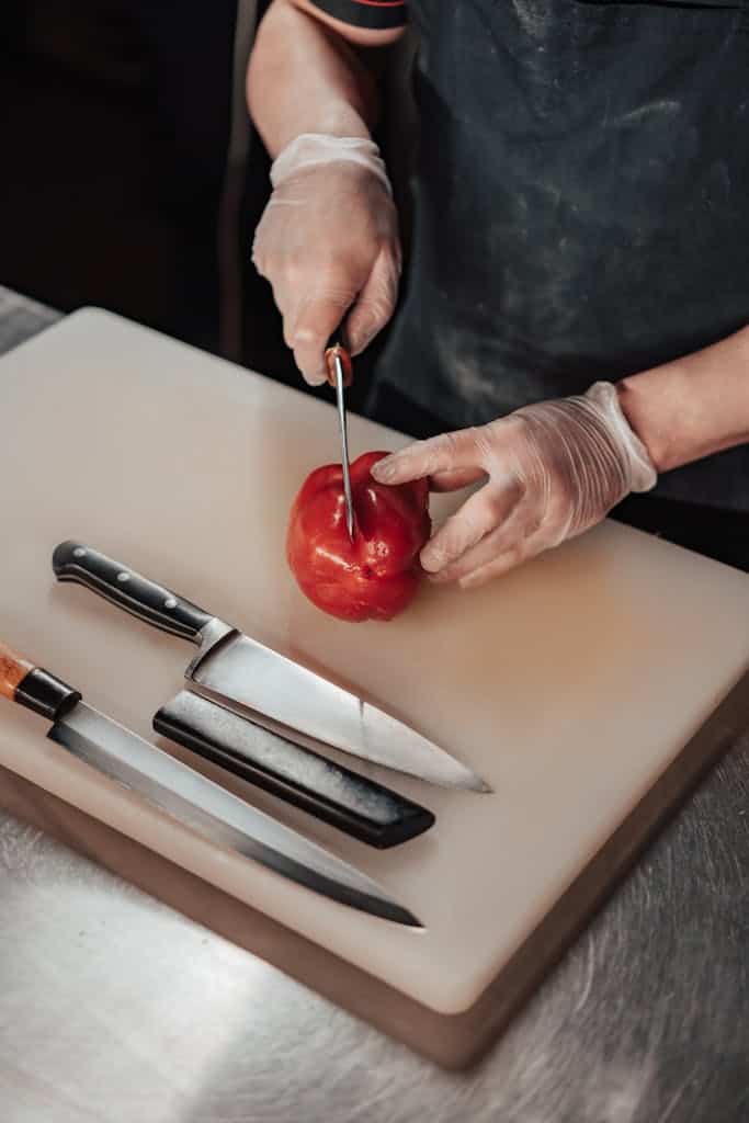 Chef's hands slicing a red bell pepper on a white cutting board with knives nearby.