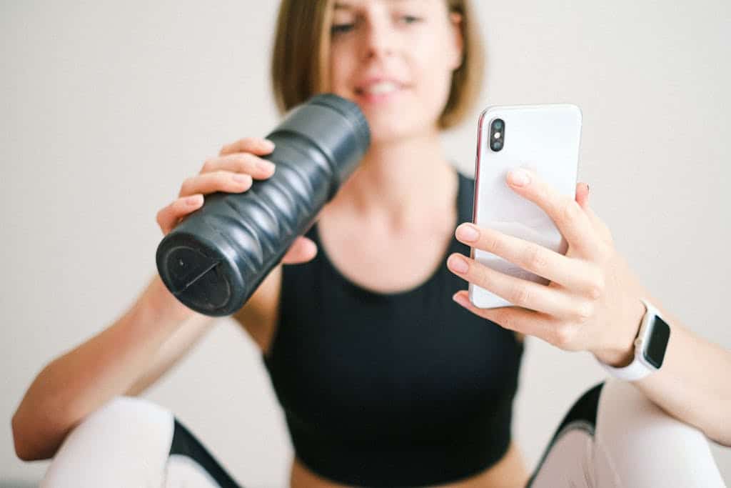Woman checks smartphone while hydrating with sports bottle during a home workout.